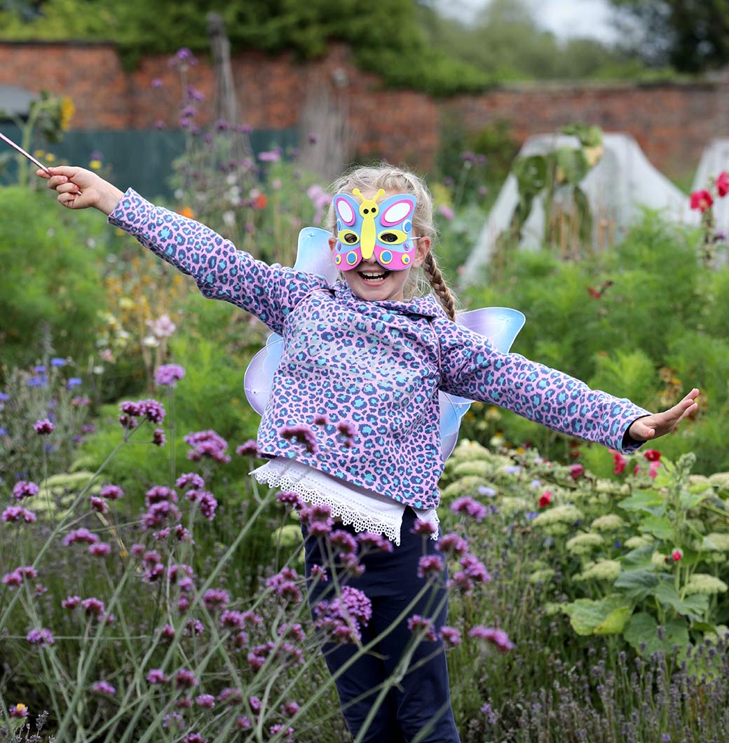 About-img-2 A child wearing a butterfly mask and wearing butterfly wings stands among flowers, arms outspread