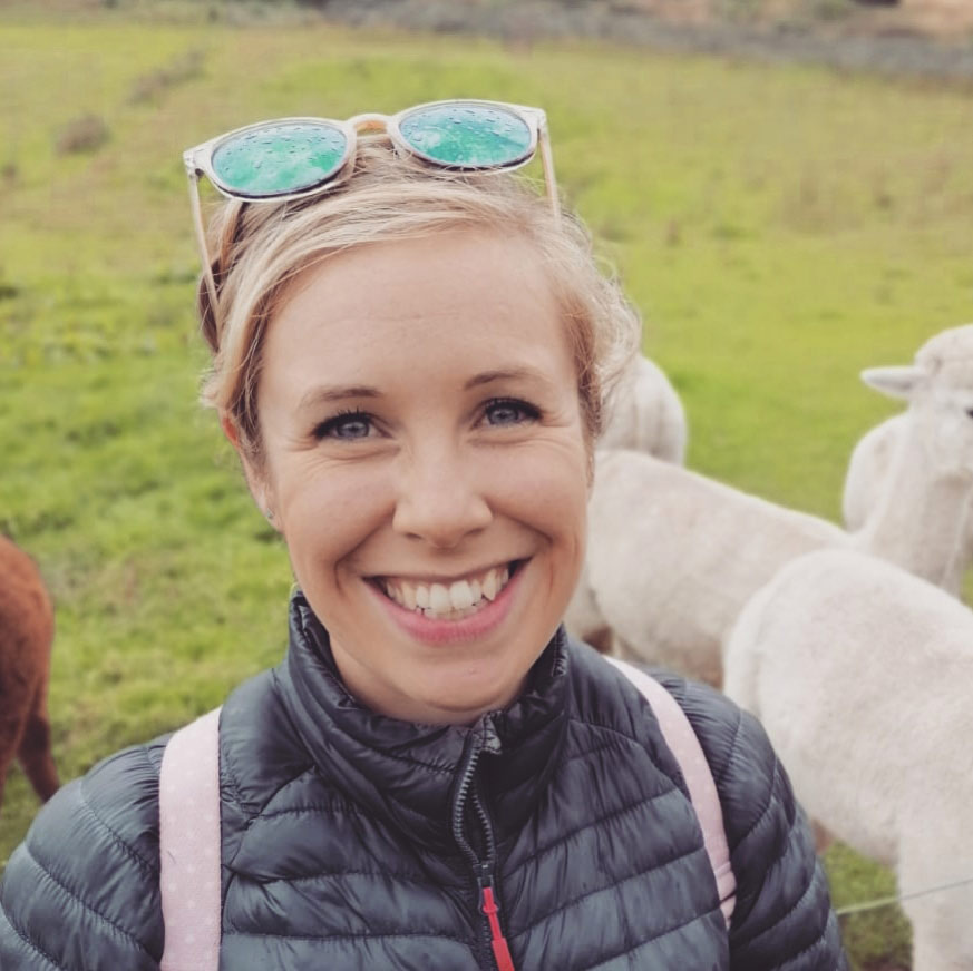 Bethanie A blonde woman wearing outdoor clothing and sunglasses on her head. There is a field of sheep behind her