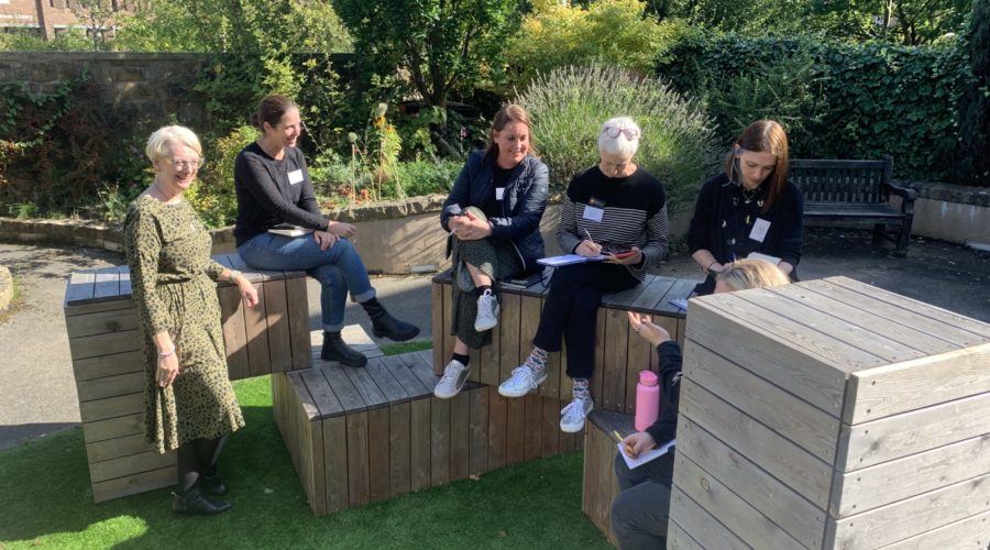A group of people sit on wooden boxes in the sunshine, chatting and making notes