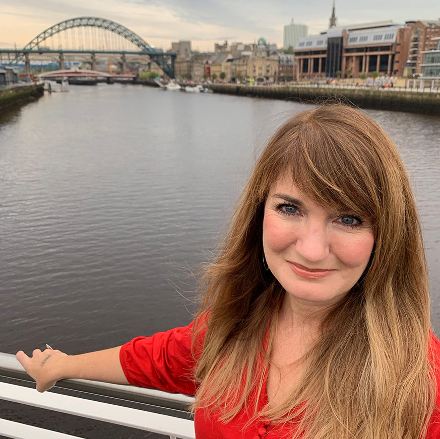 Sallyanne A woman in her fifties with long brown hair smiles. The Tyne river and bridges are behind her.