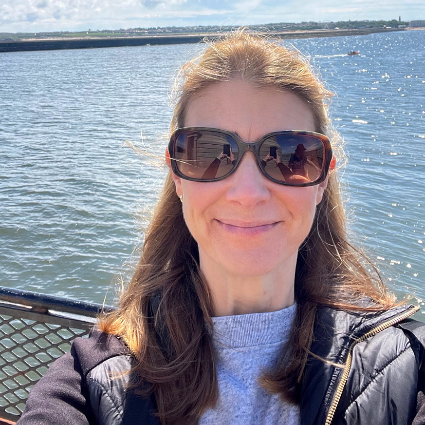 Sheryl A woman wearing sunglasses with the see behind her. She has long brown hair and is smiling.