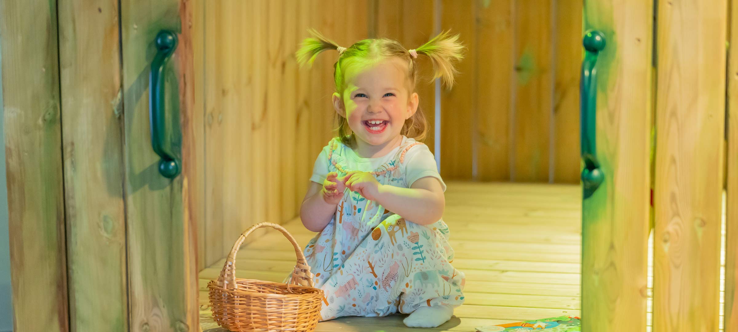 A toddler grins widely. She is inside a wooden hut and there is a wicker basket in front of her.