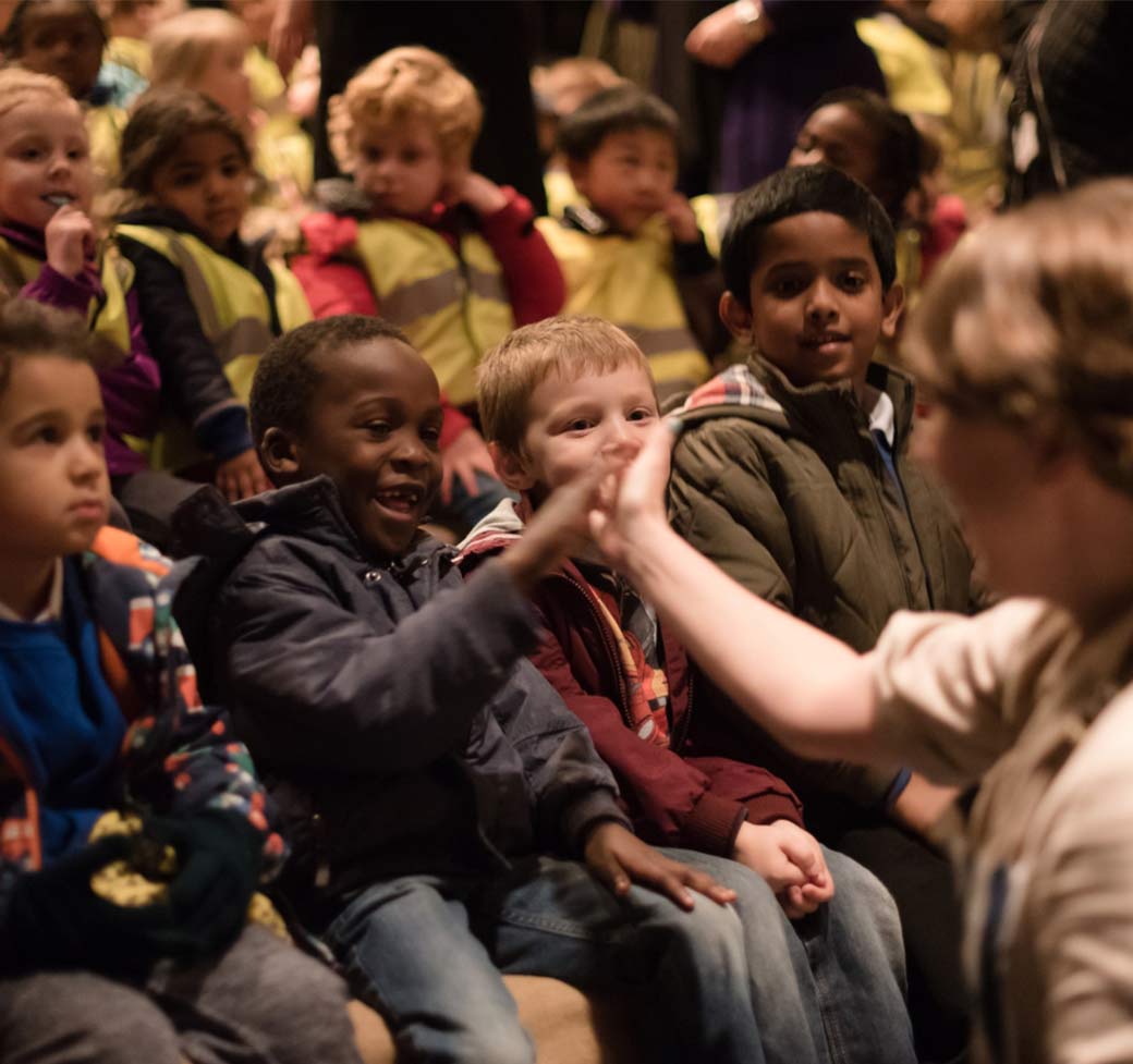 join-us-img3 A group of children sit on theatre seats. One child is high fiving an adult.