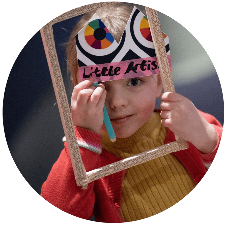A young chil peeps through a gold frame. She is wearing a handmade hat with 'Little Artists' written on it