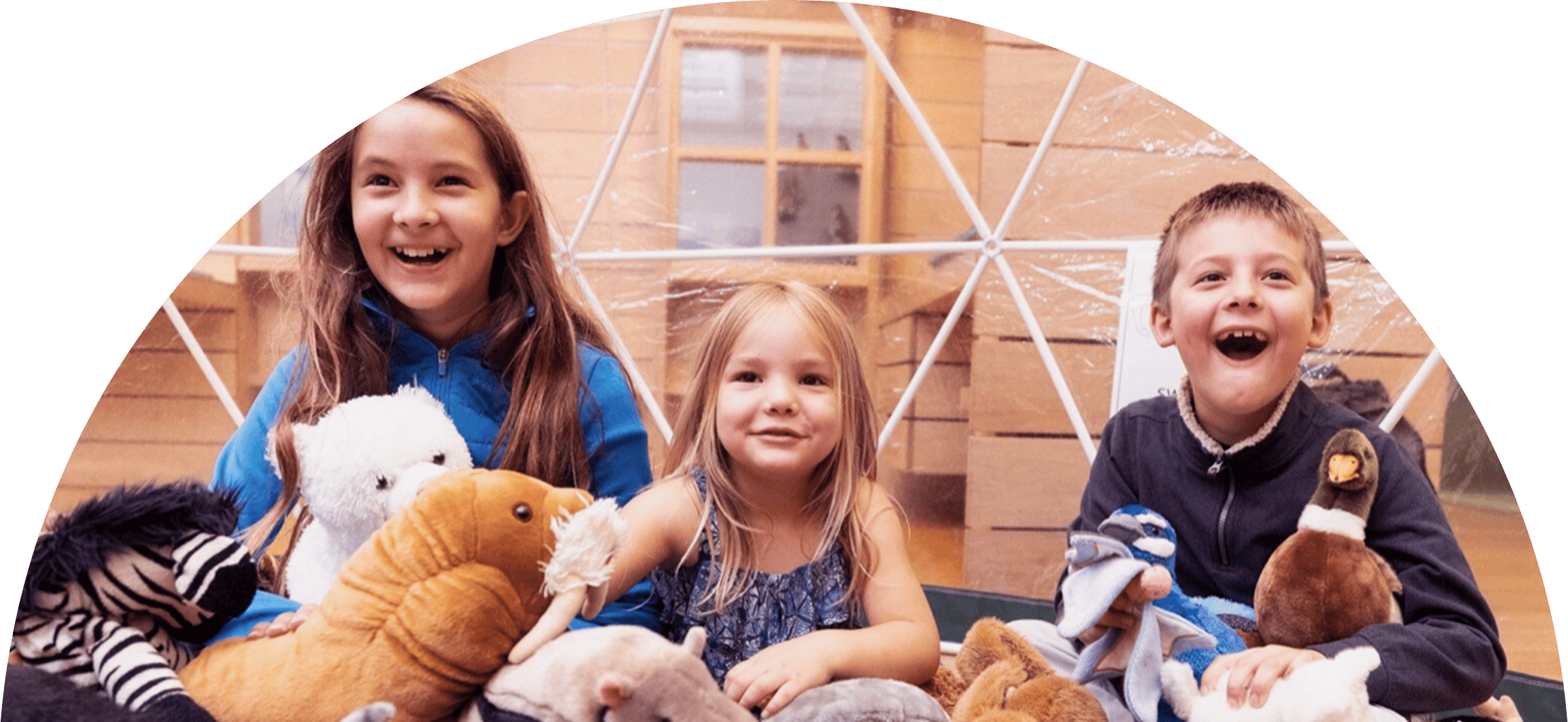 Three children sit among cuddly animal toys in a museum, grinning.