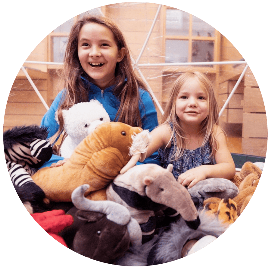 Two children sit among cuddly toys, grinning. They are inside a museum with a wooden buidling behind them.