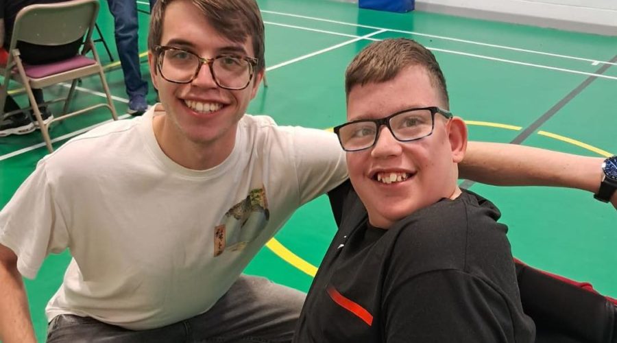 Two young people inside a gym, smiling. Bith are wearing glasses and one is in a wheelchair.