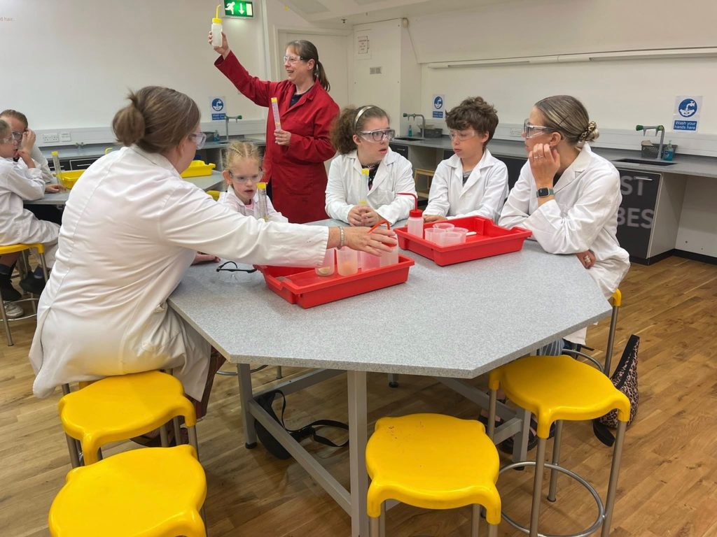 A group of adults and young people sit around a table wearing lab coats and safety glasses. A science explainer holds up a beaker in the background.